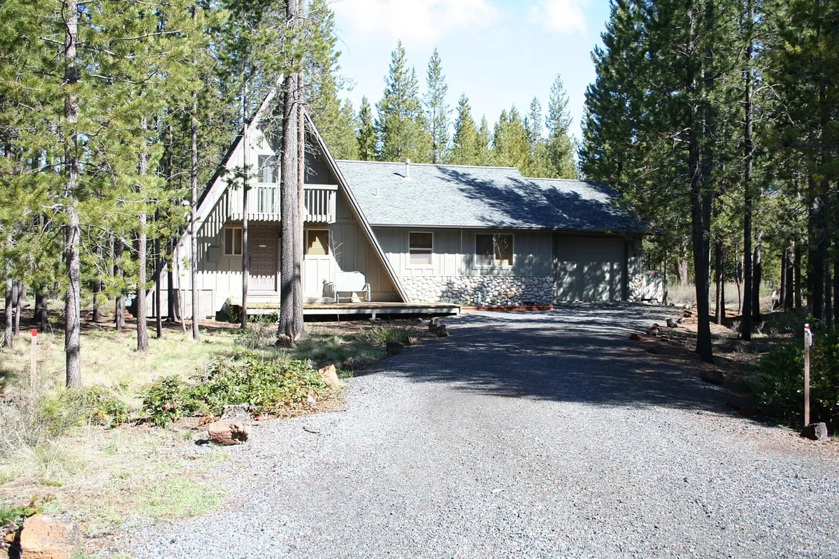 This exterior shot captures the unique A-frame structure of Trapper 11 surrounded by tall pine trees and a spacious gravel driveway. The scene evokes a sense of adventure and access to nature, inviting families to gather outside and explore the surroundings.