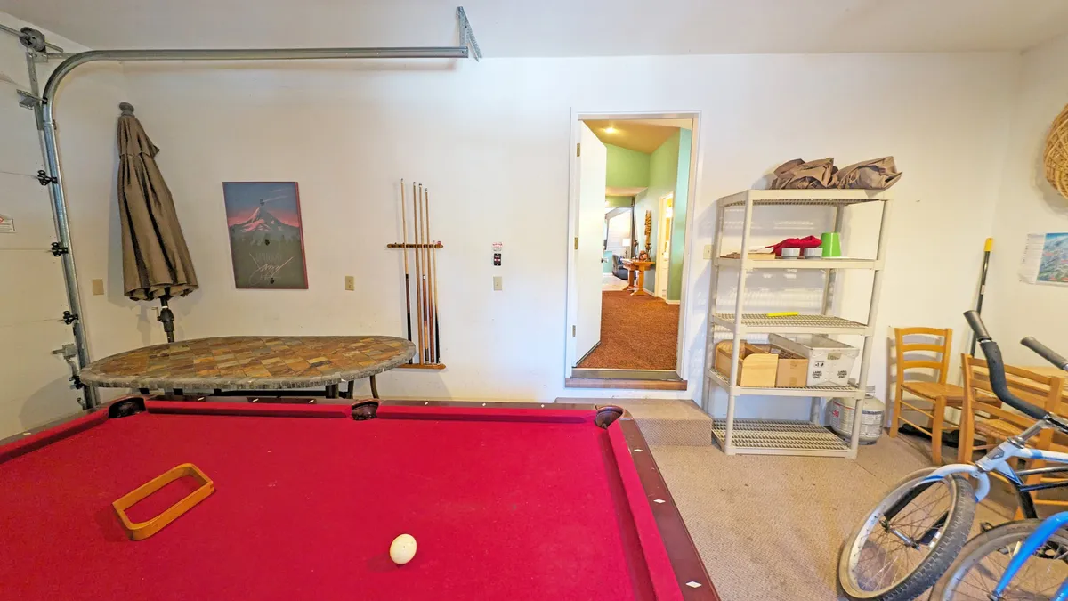 The image features another angle of the garage game room, focusing on the pool table with a bicycle and storage shelf in view. The casual layout emphasizes the relaxed and playful environment, perfect for game nights and family tournaments.