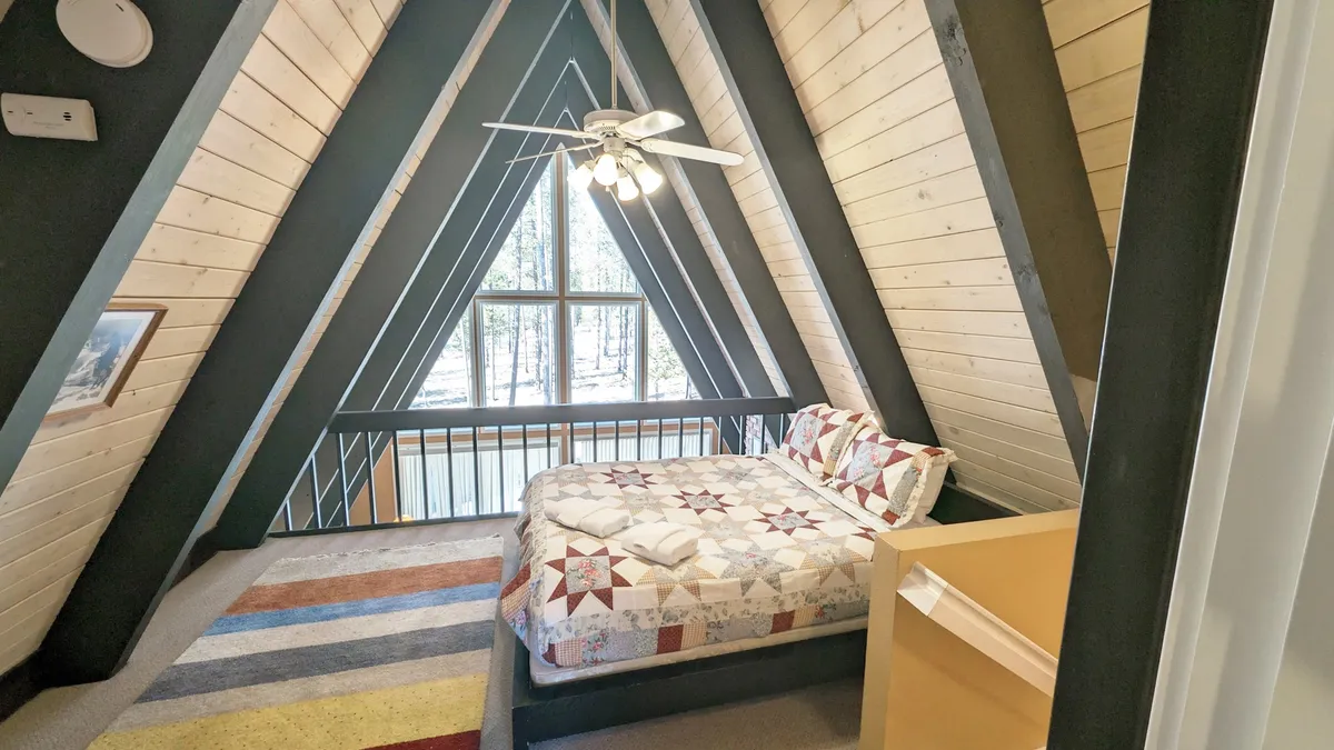 A tranquil king bedroom featuring an A-frame structure, a colorful quilt on the bed, large window with serene views, and a ceiling fan overhead. Sunlight fills the space, highlighting the wooden beams and providing a restful, airy atmosphere.