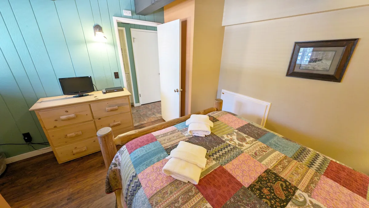 A cozy queen bedroom with a bed topped with a vibrant quilt, a wooden dresser, wall-mounted TV, and soft natural light streaming through the window. The ambiance is peaceful and welcoming—ideal for relaxation and private retreat.
