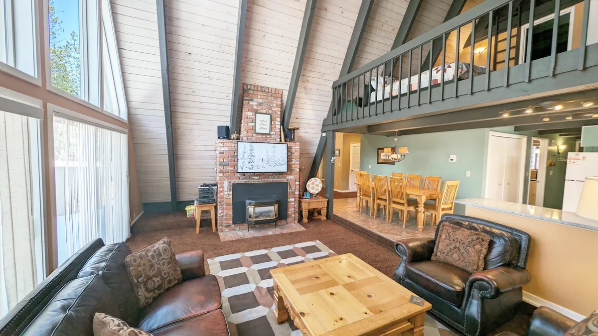 A large, welcoming room with a fireplace, expansive windows, sturdy wooden beams, and an adjoining dining area. The layout lends itself to group entertainment, whether by the fire or at the dining table, and the windows offer glimpses of the outdoors, connecting the interior to Sunriver's natural surroundings.