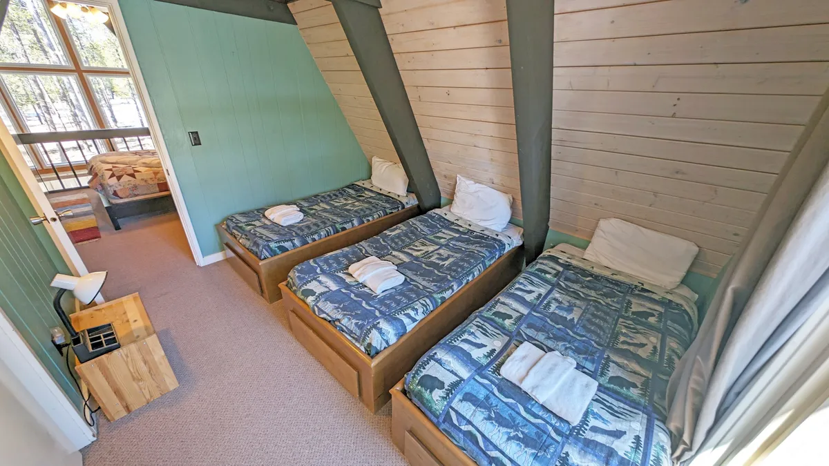 A cheerful loft space featuring three twin beds lined up beneath large windows, with light wood accents and plenty of sunlight. The area is bright and playful, perfect for younger guests or group sleepovers.