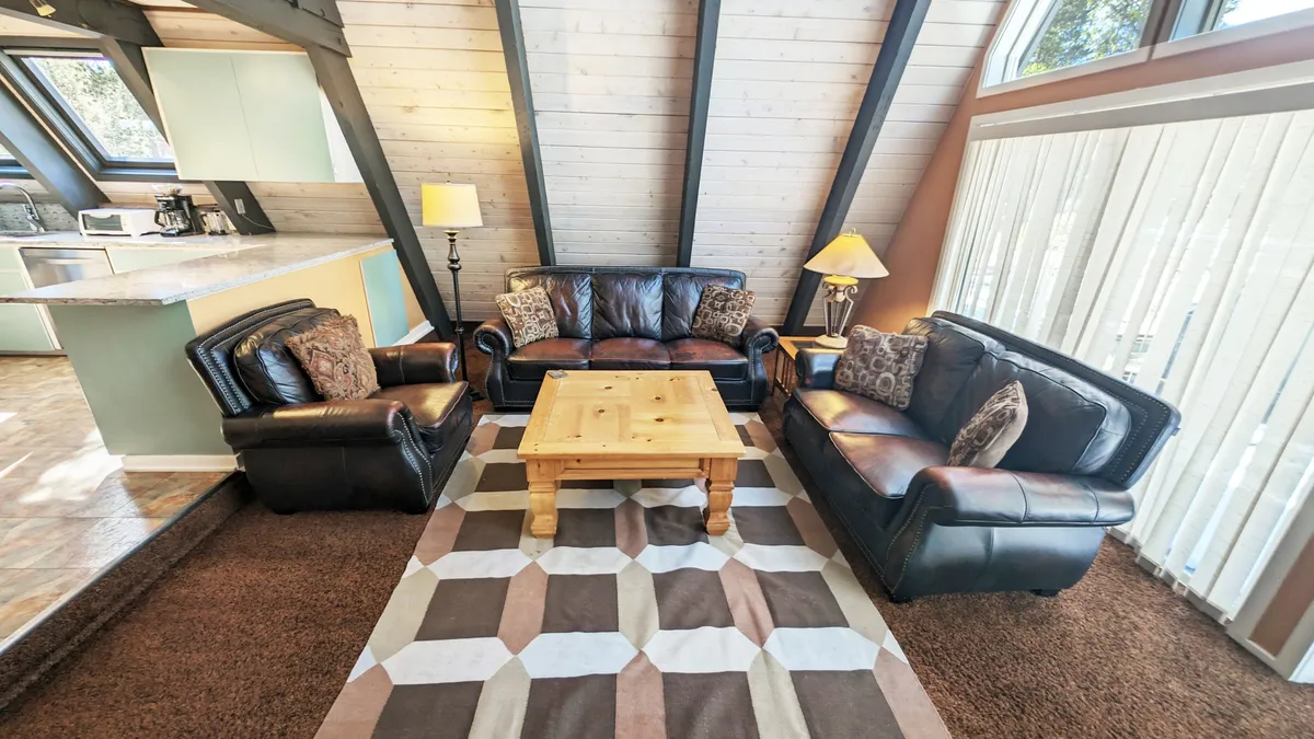 A cozy living space with leather sofas arranged around a wooden coffee table and accented by decorative pillows. Large windows flood the room with natural light, enhancing the inviting atmosphere and creating a comfortable spot for relaxing or group conversations.