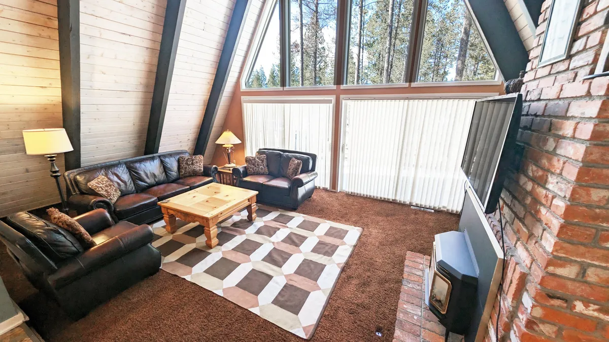 A wide view of the living and dining area featuring comfortable leather sofas, a wooden coffee table, a brick fireplace, large windows, and a textured area rug. The space is bathed in natural light, offering a sense of coziness and warmth perfect for group gatherings and game nights.