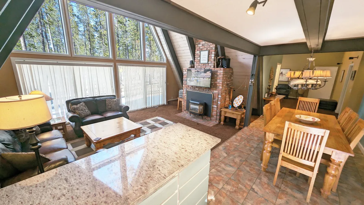 A spacious living and dining area with large windows that overlook a scenic forested landscape. The brick fireplace and comfortable seating offer a welcoming spot to unwind, while the natural light and views blur the boundaries between the indoors and the outdoors.