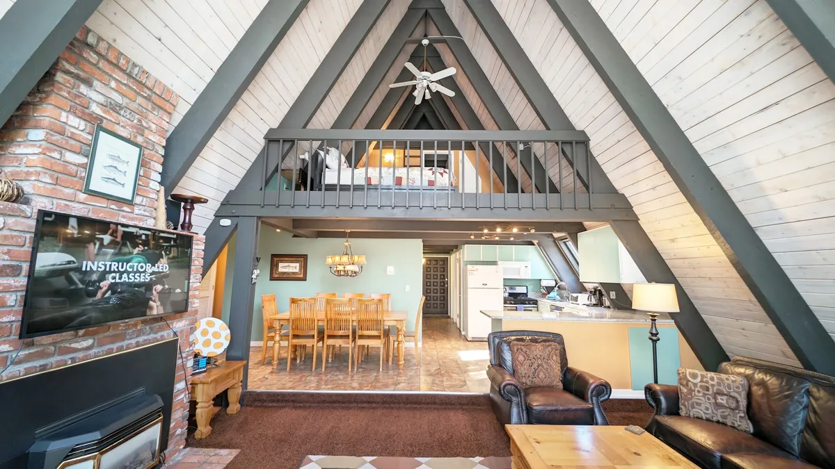 The image showcases the signature A-frame ceiling, a brick fireplace, a dining table set for gatherings, and an open loft area above. The windows draw in natural light, creating a welcoming space ideal for group activities.