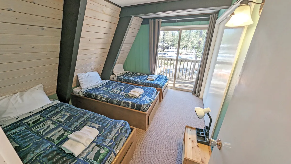 A cozy bedroom with two twin beds and a large sliding glass door that opens to a balcony. The wooden paneling and abundant natural light create a relaxed, comfortable vibe, and the balcony offers an easy transition to the outdoors.